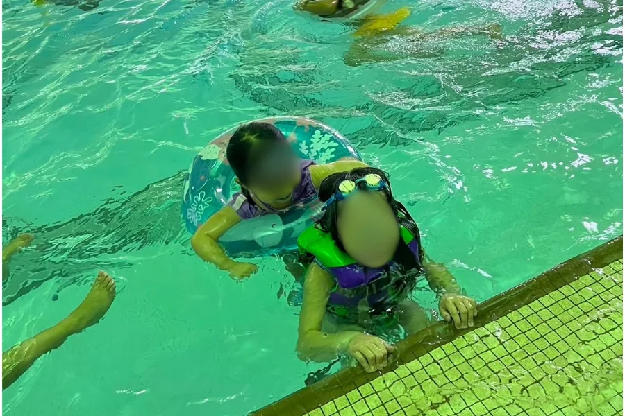 屋内プールで浮き輪をつけた子供と、ゴーグルをつけてプールサイドにつかまる子供
