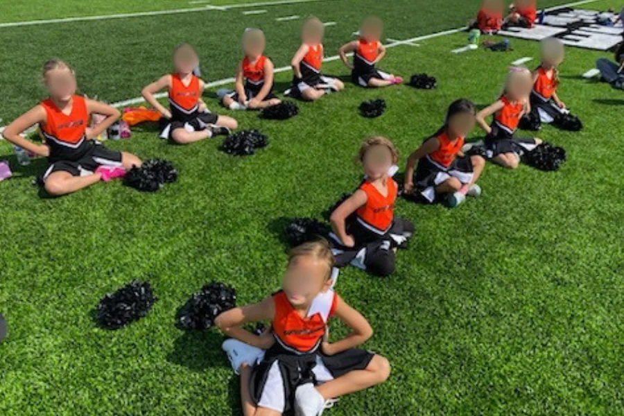 オレンジのユニフォームで芝生に座り、ポンポンを置いて休むチアの子供たち