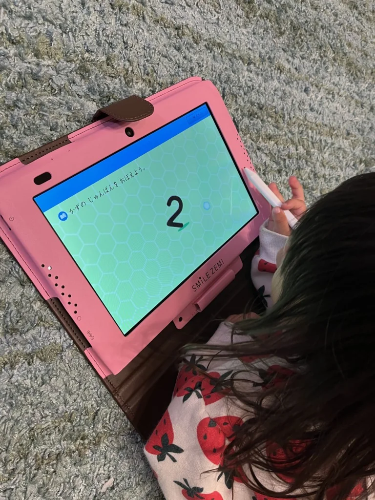 カーペットに座った子供が、スマイルゼミのタブレットにペンで数字の2を書いている。画面は「かずのじゅんばんをおぼえよう」の問題