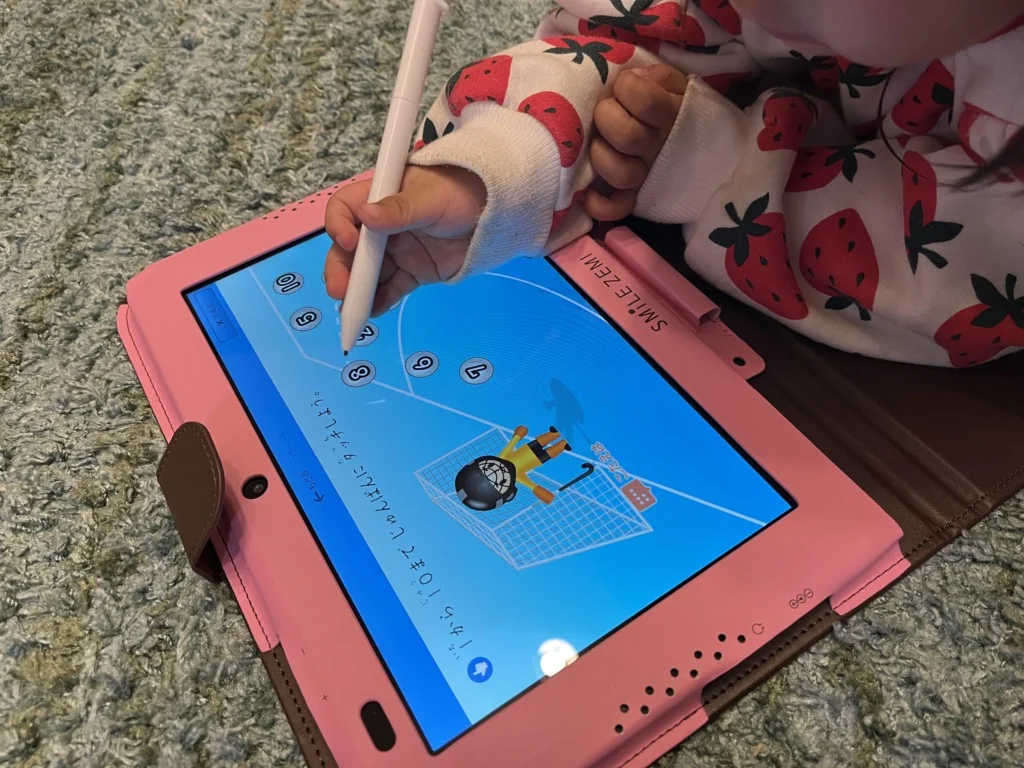 いちご柄の服を着た子供が、スマイルゼミのタブレットにペンで数字をなぞっている。画面にはサッカーゴールとキリンのイラスト